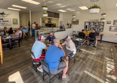 A casual cafe in Battle Lake, MN, with groups dining at tables, bright lighting, hanging plants, a bakery counter, and wall decor including framed photos and a large fish mount—experience the charm of Lake Avenue Cafe.