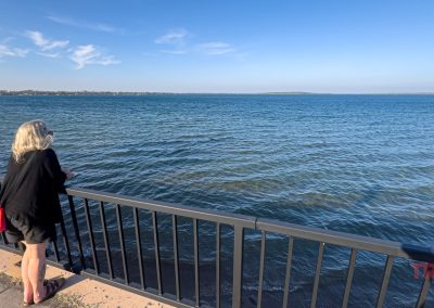 A woman with gray hair, wearing a black outfit and red bag, stands by a metal railing in Battle Lake, MN, gazing out over the vast, calm blue lake beneath a clear sky.