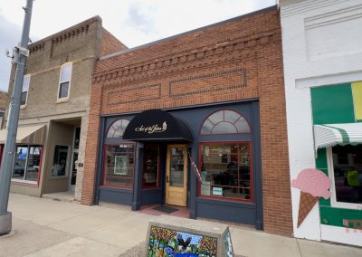 A red-brick storefront with arched windows and a black awning sits on a small-town Battle Lake, MN street, surrounded by neighboring shops. A large painted ice cream cone decorates the sidewalk near Lakes Area Arts.