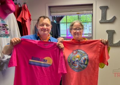 Two smiling people stand indoors holding up colorful t-shirts; the man holds a striped Battle Lake shirt, while the woman shows off an artistic outdoor scene tee. Behind them, clothing and hats from Lakes Lockerrom in MN are displayed on racks.