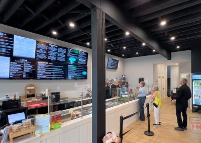 A modern café interior at Neverbetter restaurant in Battle Lake, MN, with digital menu boards, customers ordering at the counter, a glass pastry display case, and refrigerated drinks along the right wall. Black ceiling beams and bright lighting create an inviting atmosphere.
