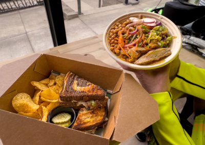 A person holds a bowl of salad with beans, chicken, and vegetables. Next to it is a box with potato chips, pickles, and a grilled sandwich on a wooden table by a window at Neverbetter restaurant in Battle Lake, MN.