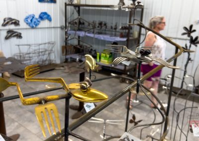 Metal sculptures from repurposed forks and spoons, crafted by Pauls Metal Petals, are displayed in a bright room in Battle Lake, MN, as a woman observes the unique art in the background.