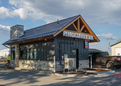 A small modern drive-thru coffee shop in Battle Lake, MN, with a black truck parked at the window and a cyclist waiting nearby. The building features a sloped metal roof, stone accents, and a sign reading Perks Coffee & Tea.