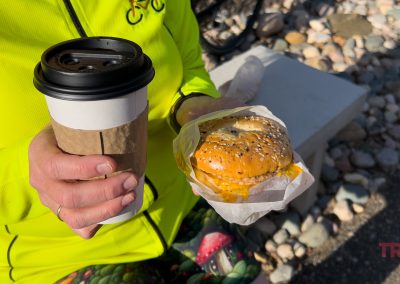 A person in a bright yellow jacket holds a Perks Coffee cup in one hand and a cheesy bagel sandwich in the other, enjoying the sunlight near Battle Lake, MN, with shadows playing across the rocky landscape.