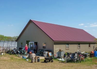 A tan metal building with a red roof sits under a blue sky at Pixie Woods Antiques in Battle Lake, MN. Bikes, chairs, and tools are scattered outside, while two people stand near the entrance surrounded by trees and equipment.
