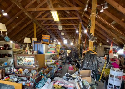 Interior of Pixie Woods Antiques in Battle Lake, MN—a thrift shop with shelves full of lamps, dishes, figurines, and clothes, all beneath a wooden vaulted ceiling with exposed beams.