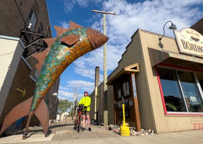 A cyclist in a bright yellow jacket stands beside Rusty the Fish, a large, colorful sculpture in an alleyway between brick buildings in Battle Lake, MN, one with a “Boathouse” sign, beneath a blue sky dotted with clouds.