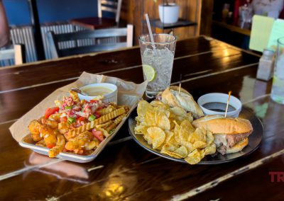 A wooden table at The Boathouse restaurant in Battle Lake, MN, features crinkle-cut fries topped with tomatoes, cheese, and herbs, a burger with chips, two dipping sauces, and drinks with lime slices.