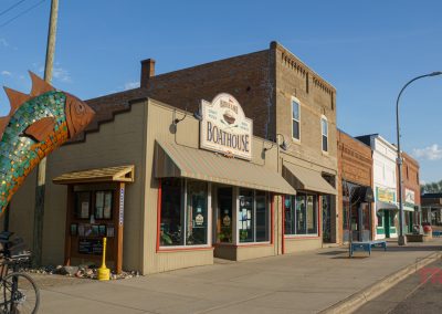A small-town street in Battle Lake, MN, with brick buildings including The Boathouse restaurant. A large, colorful fish sculpture stands outside, and bicycles are parked by the sidewalk on a sunny day.