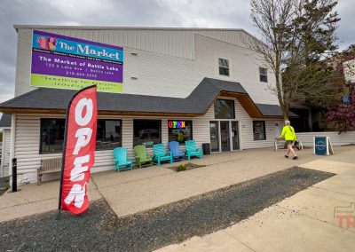 A man in a yellow jacket walks past a storefront with a large “OPEN” flag, colorful chairs, and a sign for The Market of Battle Lake in Battle Lake, MN on a cloudy day.