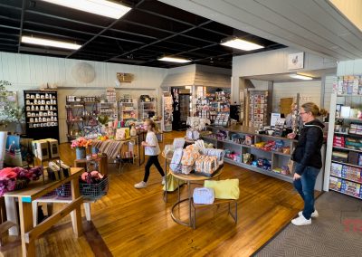 A bright, inviting gift shop in Battle Lake, MN, The Market store features shelves of books, games, home decor, and more. Three people—including a child—browse the merchandise as wooden floors and display tables add warmth to the space.