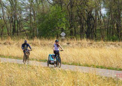 Two people ride bikes along the Glendalough Trail through tall, dry grass with trees in the background. One cyclist pulls a blue child carrier. It is a sunny day at Glendalough State Park, and both riders are wearing helmets.