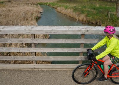 A person wearing a bright yellow jacket and pink helmet rides a red bike across a wooden bridge over a small creek lined with dry reeds along the Glendalough Trail in Glendalough State Park.
