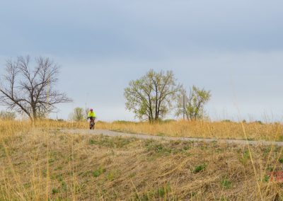 A cyclist wearing a bright neon jacket rides along the Glendalough Trail, a paved path winding through a grassy field dotted with leafless and green trees under a cloudy sky in Glendalough State Park.