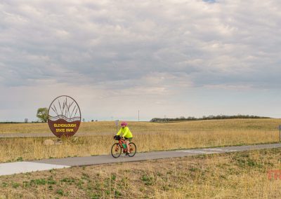 A cyclist in a bright yellow jacket and pink helmet rides along the Glendalough Trail, passing the sign for Glendalough State Park, surrounded by dry grass under a cloudy sky.