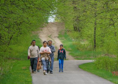 A family of five walks along the Glendalough Trail at Glendalough State Park, surrounded by lush green trees and grass on a cloudy day. The group includes three adults and two children, strolling together in casual clothing.