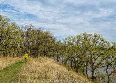 A person in a bright yellow shirt jogs along a grassy trail on a hill overlooking a lake at Glendalough State Park, with trees and a partly cloudy sky in the background.