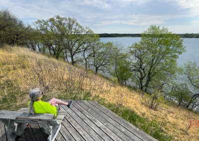 A person with long gray hair, wearing a bright green shirt, sits on a wooden bench overlooking the lake at Glendalough State Park, surrounded by trees and grassy hills under a partly cloudy sky.