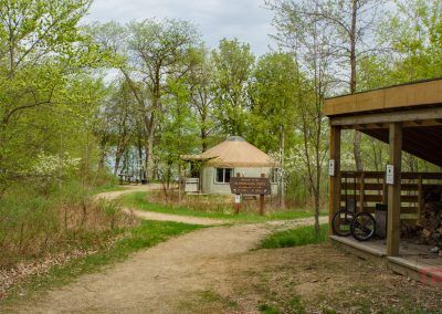 A dirt trail winds through green trees toward a round yurt structure in Glendalough State Park. A wooden shelter with stacked firewood and two bikes is on the right, with a sign and a glimpse of water visible in the background.