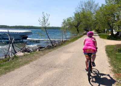 A cyclist in a pink outfit rides a bike along a lakeside path with trees and a docked boat nearby. A sign reads “Speed Limit 10.” The weather is sunny with blue skies and the water is slightly choppy.