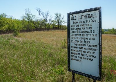 A historical sign in a grassy field along the Otter Trail Scenic Byway describes the settlement of Old Clitherall by a group of Latter-Day Saints. Trees and blue sky are visible.