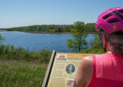 A woman in a pink helmet and sleeveless top reads an informational sign about Clitherall Lake along the Otter Trail Scenic Byway, overlooking a scenic view of the lake, trees, and grassy shoreline on a sunny day.