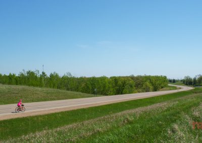 A person in a pink outfit rides a bicycle along a paved road, the Otter Trail Scenic Byway that curves through green grassy hills and trees under a clear blue sky. The scene is calm and open, with lots of natural landscape.