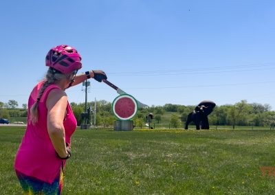 A woman in bright cycling gear stands on grass in Vining, MN, gazing at a large Nyberg Sculpture watermelon slice and spoon, with a black elephant statue in the background under a clear blue sky.