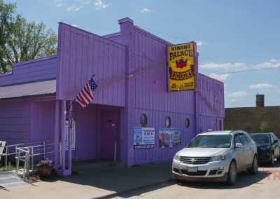 A bright purple liquor store with an American flag, a yellow “Vining Palace Liquors” sign, and several cars parked in front on a sunny day in a small town.