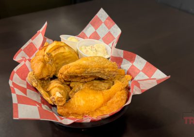 A basket lined with red and white checkered paper holds crispy fried chicken, potato wedges, and two small cups of dipping sauces on a dark table at Purple Palace Restaurant.