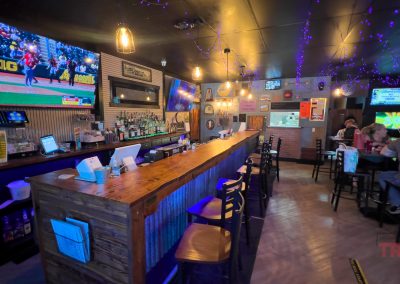A cozy sports bar, Purple Palace Restaurant, features a long wooden counter, bar stools, several TVs showing sports, purple string lights on the ceiling, and a few people dining at tables in the background.
