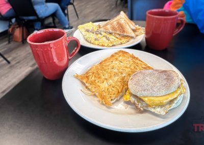 A breakfast table at 8th Ave Diner in Pipestone, MN displays two red mugs, scrambled eggs with toast, and a plate with a breakfast sandwich, melted cheese, and hash browns. Two people in jeans sit in the background.