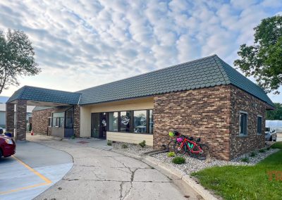 A one-story brick building, 8th Ave Diner, with green roof tiles, large front windows, and bicycles parked outside sits in Pipestone MN. Cars fill the lot and trees frame a cloudy sky.