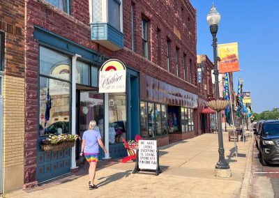 A woman in a tie-dye skirt walks on a sunny sidewalk past a brick building that houses Clothier By Dawn boutique with shops and a sign that reads, “We have loans, evening closing soon,” in a small downtown area with parked cars and street banners.
