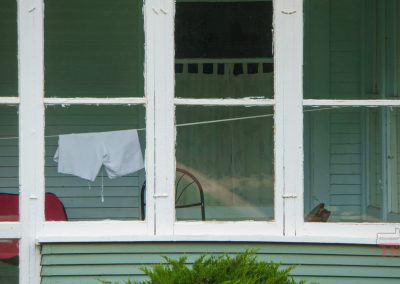 A white shirt pair of bloomers hangs on a clothesline inside a porch with light blue-green walls and white framed windows; a red chair and a wooden chair are visible in the background.