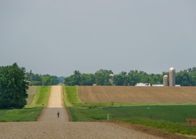 A lone cyclist travels down a long, straight gravel road through rural farmland, bordered by fields and trees. Silos and a barn are visible in the distance under a hazy sky.