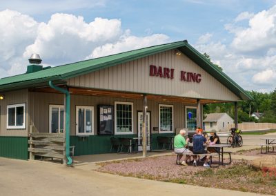 A small building with a green roof and Pipestone Dari King sign, with a few people sitting at an outdoor table. Bicycles are parked nearby, and trees and clouds are in the background.