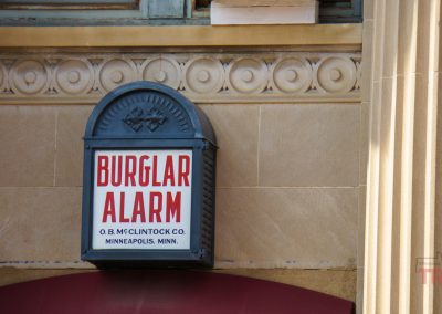 A vintage metal burglar alarm box with red and black text is mounted on a tan brick building wall, featured along the Pipestone History Walk. The alarm reads Burglar Alarm, O.B. McClintock Co., Minneapolis, Minn.
