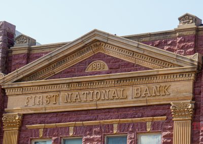The image shows the upper facade of a historic brick building in Pipestone MN, with a stone pediment inscribed FIRST NATIONAL BANK and the year 1898 above it—an ornate highlight along the city’s History Walk.
