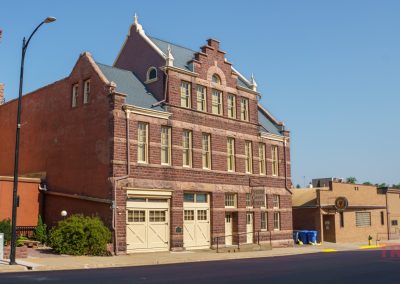 A historic red stone building with ornate windows and a steep gabled roof sits on a sunny street corner in Pipestone MN, perfect for a History Walk. Across the way stands a newer tan brick structure under the clear blue sky.