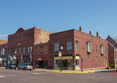 A sunny street scene in Pipestone, MN with historic red brick buildings, parked cars, and colorful flags along the History Walk. Clear blue sky overhead, with sidewalks and streetlights lining the road in this charming downtown area.