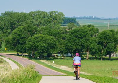 A cyclist wearing a pink helmet and colorful shorts rides along the Indian Lakes Trail in Pipestone MN, following a paved path through green fields, trees, and a winding road beneath a clear sky.