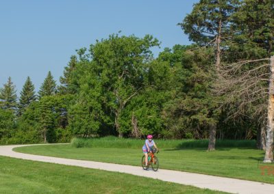 A person wearing a pink helmet rides a bicycle along the Indian Lakes Trail, a paved path winding through a grassy park with tall green trees under the clear blue sky of Pipestone MN.