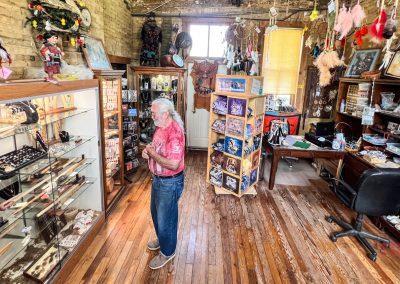 An elderly man with long white hair stands in a small shop with wooden floors, surrounded by glass display cases of jewelry, dreamcatchers, and Indigenous art—an inviting space curated by Bud Johnston of the Pipestone Keepers.