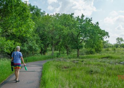 A person with gray hair walks on a paved trail through a green, grassy park at Pipestone National Monument, carrying water bottles. Tall trees line the path under a partly cloudy sky.