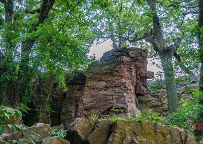 Large reddish-brown rock formations, like those at Pipestone National Monument, rise amid dense green trees and foliage on a cloudy day, with patches of moss and lichen adding character—some rocks even resemble an old stone face.