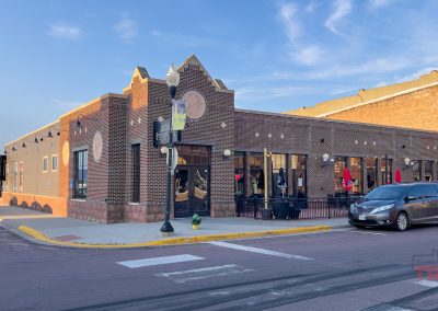 A brick Stonehouse Restaurant with outdoor seating on a corner in Pipestone, MN, a parked car nearby, streetlamp, and clear blue sky in the background. The building features large windows and decorative round plaques on the facade.