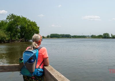 A woman with gray hair and a blue backpack looks out over a calm, wide river at Split Rock Creek State Park. Trees line the riverbank, and wind turbines are visible in the distance under a partly cloudy sky.