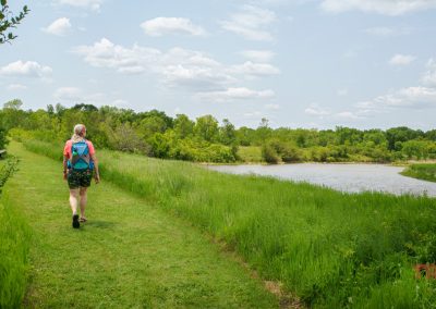 A person with a blue backpack walks on a grassy trail alongside a river at Split Rock Creek State Park, surrounded by green trees and under a partly cloudy sky.
